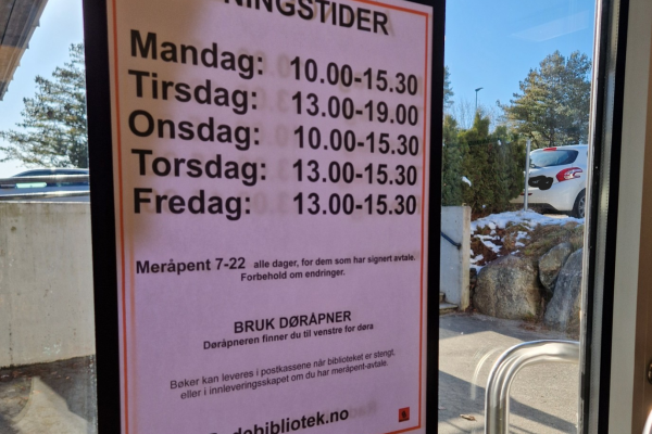 Bilde viser plakat med åpningstider på inngangsdør til biblioteket sett fra innsiden