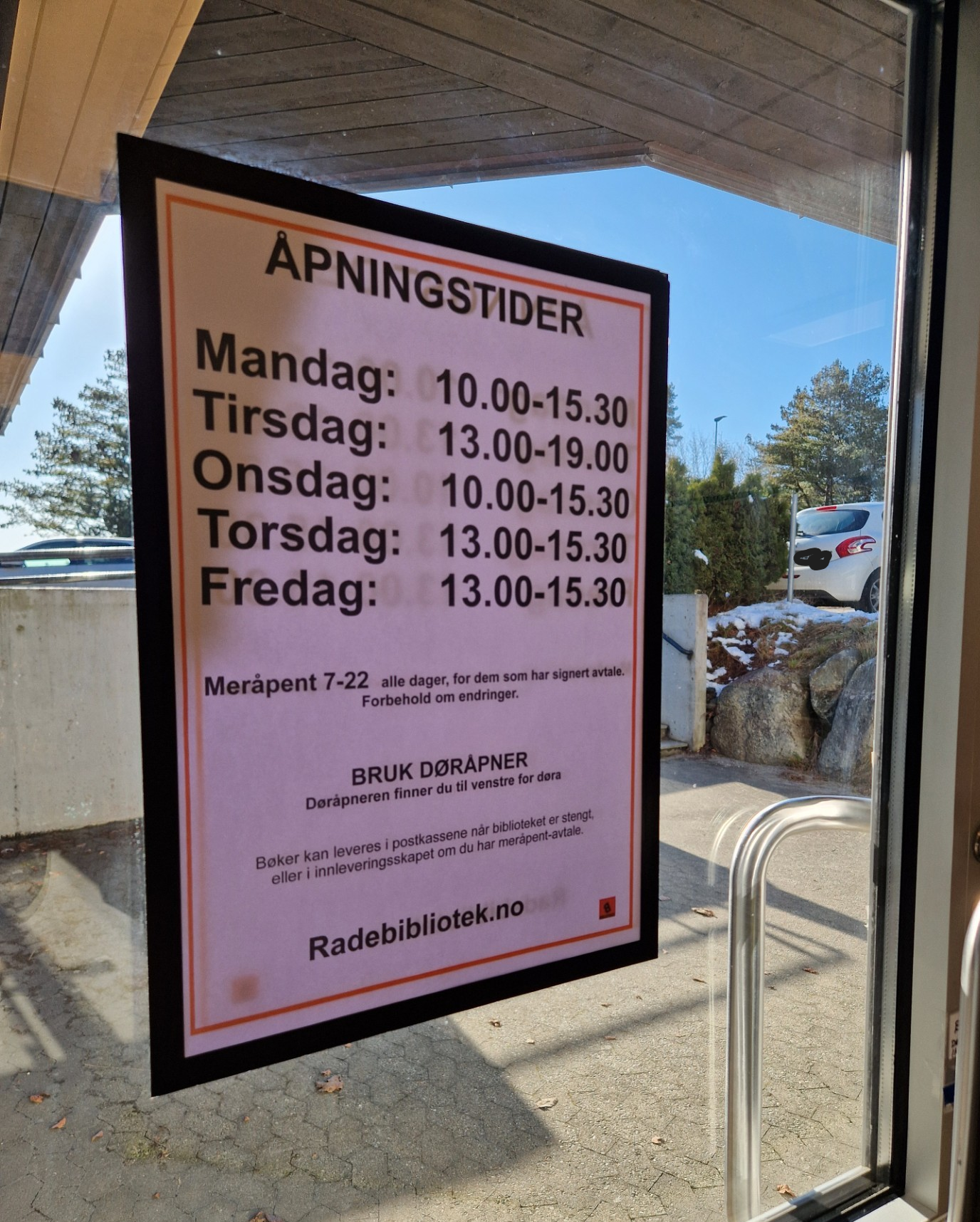 Bilde viser plakat med åpningstider på inngangsdør til biblioteket sett fra innsiden