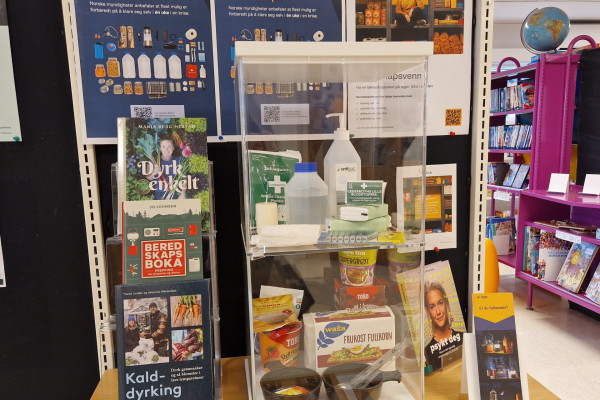 Utstilling i anledning egenberedskapsuka. Monter på bord ved siden av bøker og med plakater om egenberedskap på tavle bak. I monteren er utstilt knekkebrød, plaster og andre ting det er lurt å ha i boden eller kjelleren